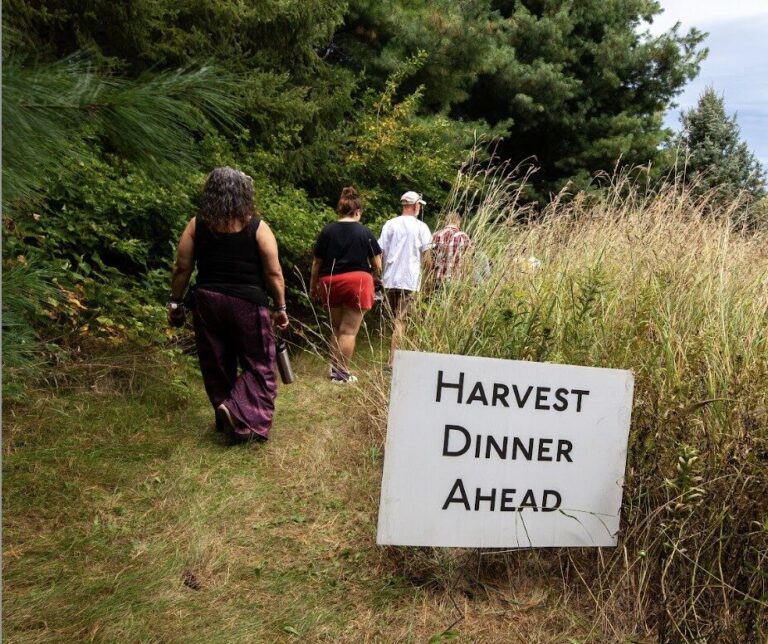 A line of people walking a grassy path through trees with a sign in the foreground that says, "Harvest Dinner Ahead."